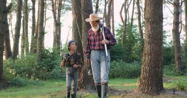 Gros plan d'attrayant calme respecté grand-père barbu chapeau qui va à la pêche avec son beau petit-fils intéressé, en passant par le parc vert — Video