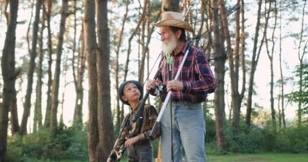 Gros plan de séduisant homme âgé barbu respecté chapeau et son petit-fils beau qui parle tout en allant pêcher avec des cannes et filet de pêche à travers le parc — Video