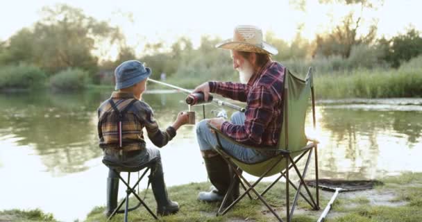 Bello positivo vecchio nonno barbuto riempiendo la tazza di tè il suo bel nipotino durante la pesca sul laghetto, al rallentatore — Video Stock