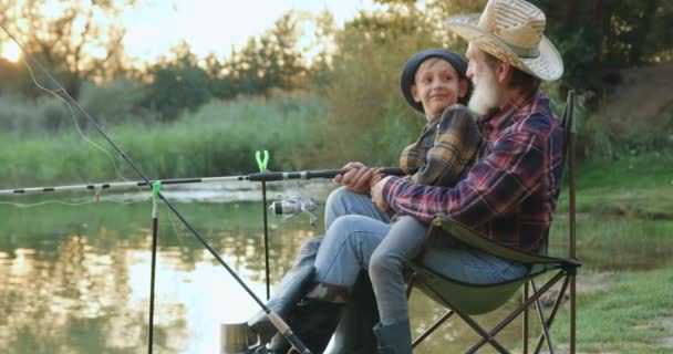 Vista lateral de bonito sorrindo menino pequeno feliz que sentado em granndfathers joelhos enquanto eles pescando com varas na lagoa no verão — Vídeo de Stock