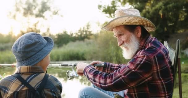 Summer family recreation concept where attractive mature bearded granddad telling fishing sagacity to his interested small grandson while they fishing together on the pond — Stock Video