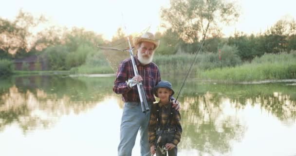 Vorderansicht des gut aussehenden lächelnden fröhlichen bärtigen Großvaters und des 10-jährigen Enkels, die Angelruten in der Hand halten und am Ufer der Teiche vor der Kamera posieren