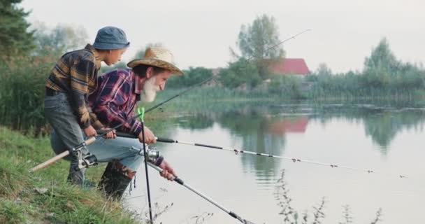 Likable positivo anziano barbuto pescatore pesca insieme con il suo 10-aged gioioso bel nipote utilizzando canne da pesca e rete sulla riva stagni durante il giorno in estate — Video Stock