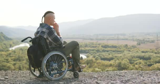 Πλευρική άποψη του όμορφος ήρεμος συμπυκνωμένος άνθρωπος με μικρή αλογοουρά που κάθεται σε αναπηρική καρέκλα στο λόφο και προσεύχεται — Αρχείο Βίντεο