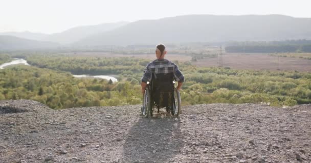 Невідомий сидячий молодий чоловік з надзвичайним зачіскою сидить у інвалідному кріслі і дивиться на завоювану природу навколо пагорба, вид ззаду — стокове відео