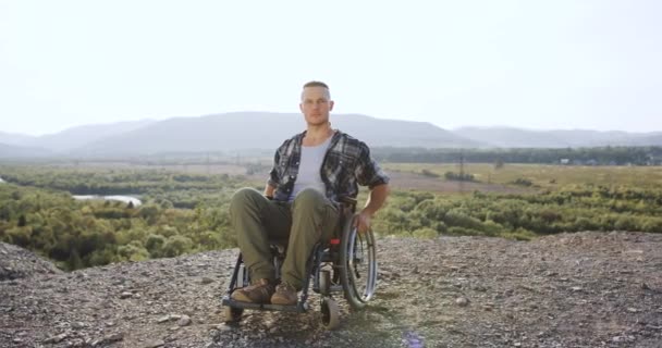 Tampan serius pria 30-tahun cacat di kursi roda Melihat kamera di mount pada latar belakang pemandangan indah — Stok Video