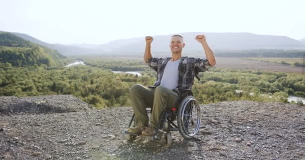 Bonito positivo feliz jovem deficiente em cadeira de rodas levantou as mãos sobre o monte em rodeado natureza maravilhosa, vista frontal — Vídeo de Stock