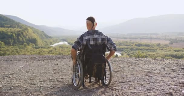 Visão traseira do jovem incapacitado em cadeira de rodas que cavalgando até o final da colina para olhar ao redor sobre as paisagens pitorescas durante o dia — Vídeo de Stock