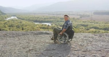 Tekerlekli sandalyedeki çekici, genç, engelli adamın yan görüntüsü. Tepenin tepesinde manzaraya bakıyor.