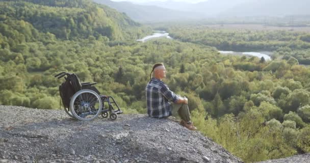 Beau jeune homme handicapé chanceux et souriant assis sur la pierre au sommet de la colline et respirant sur la poitrine pleine profitant de la beauté de la nature pittoresque, vue latérale — Video