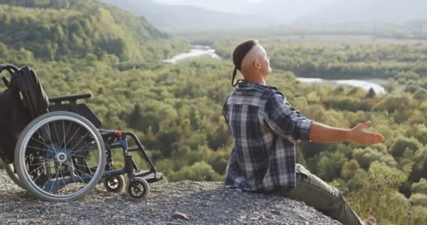 산 정상에 있는 그의 휠체어 근처에 앉아 그림같은 자연 속에서 태양 아래서 그의 얼굴을 대체하는 행복 한 젊은 남자 다운 모습 — 비디오