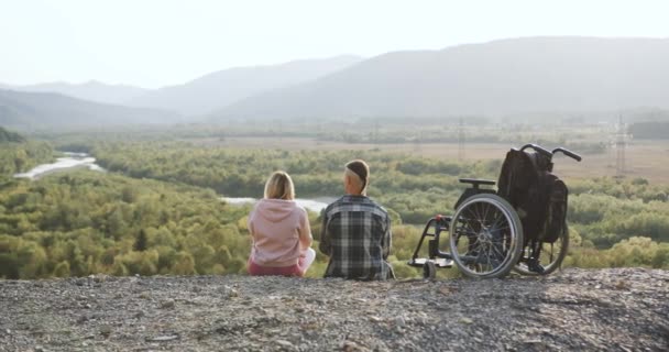 Концепция семейного отдыха, где молодая счастливая любящая пара, где один из них инвалид сидит на холме и наслаждается окружающими живописными пейзажами — стоковое видео