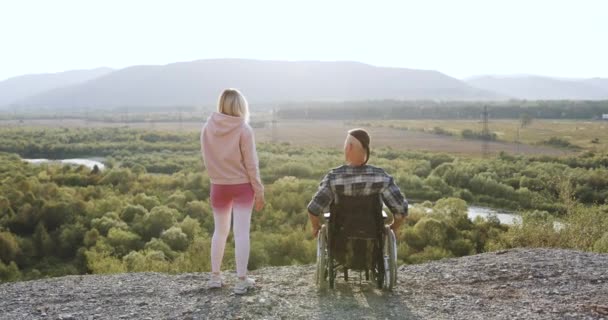 Обратный вид привлекательного расслабленного инвалида в инвалидной коляске и его спортивной жены, которая стоит на вершине холма и держит их за руки, наблюдая за красивой природой вокруг — стоковое видео
