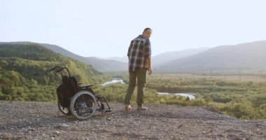 Yakışıklı, uzun boylu, güçsüz bir adamın arka görünüşü. Tekerlekli sandalyeden kalkıp kollarını uzatan ve güneş ışığı altında özgürlüğün tadını çıkaran bir adam.