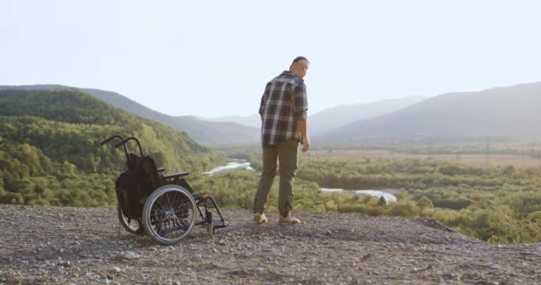 Bakspeilet på en kjekk, høy, midlertidig arbeidsudyktig mann som reiser seg fra rullestol på hesten, strekker armene og nyter perfekte frihetsfølelser under sollys – stockvideo