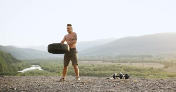 Utsikt til kjekk, muskuløs, skjorteløs høy, ung mann i brun shorts som gjør kraftøvelser med dekk fra hjul på hesten med vakker natur rundt – stockvideo