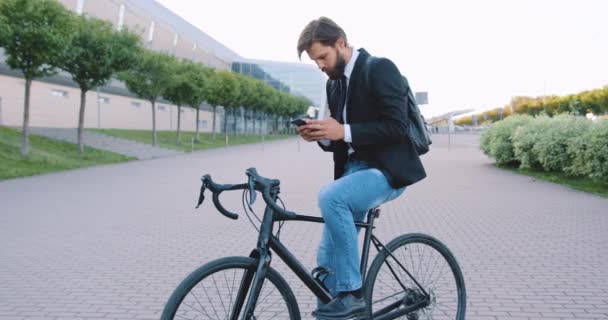 Vonzó elegáns, fiatal szakállas férfi ül kerékpáron megállás közben a gyalogos útvonal a város épület hátterében nappal, elölnézet — Stock videók