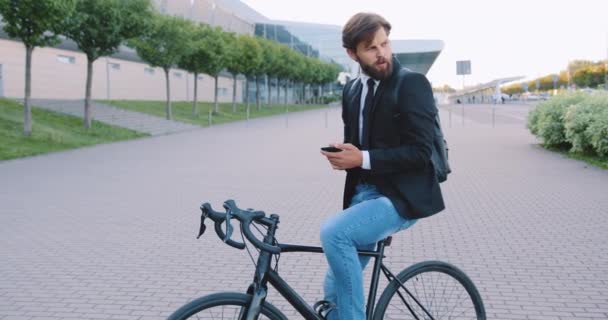 Портрет приємного серйозного молодого бородатого офісного працівника, який сидить на своєму велосипеді і використовує телефон на великому міському будівельному тлі — стокове відео