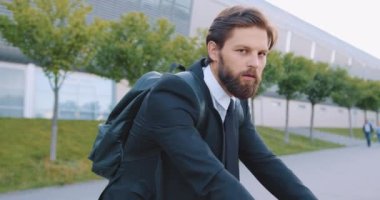 Modern şehir binasının arka planında bisiklet üzerinde otururken kameraya poz veren yakışıklı, şık, havalı, sakallı genç adam.