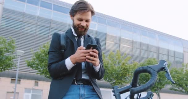 Widok z przodu uśmiechnięty przystojny, stylowy brodaty mężczyzna, który siedzi na rowerze i przegląda śmieszne filmy na telefonie w pobliżu pięknego nowoczesnego budynku miasta — Wideo stockowe