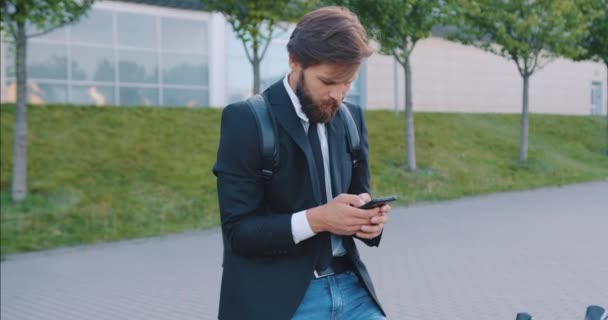 Bonne humeur positive heureux jeune barbu élégant gars arrêter sur son vélo et écriture messege sur son smartphone près de beau bâtiment urbain moderne — Video