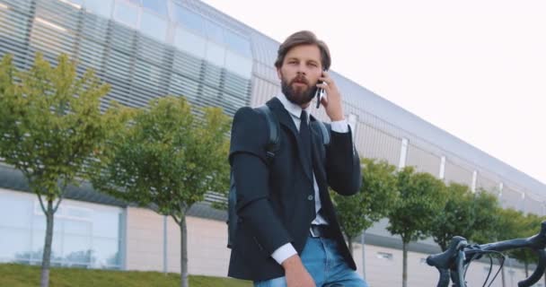 Гарний впевнений позитивний молодий бородатий чоловік має мобільну розмову на відкритому повітрі, сидячи на своєму велосипеді біля великої міської будівлі — стокове відео