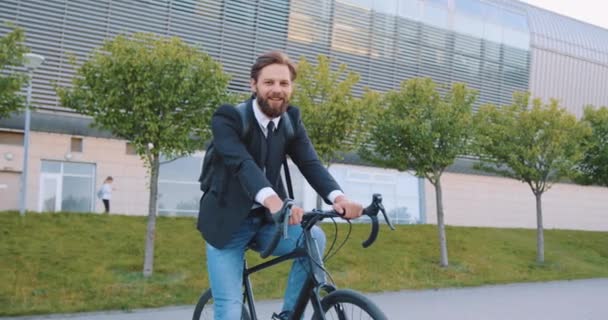 Atraente sorridente despreocupado jovem barbudo em roupas elegantes sentado em sua bicicleta e olhando para a câmera perto de edifício urbano grande moderno — Vídeo de Stock