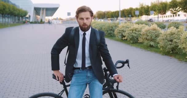 Close up de homem elegante moderno bonito sério com barba que sentado em sua bicicleta no caminho pedestre perto de grande construção urbana e olhando para a câmera — Vídeo de Stock