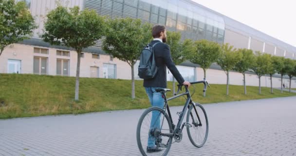 Güzel modern şehir binasının yanındaki kaldırımlı caddede bisikletiyle yürüyen ceketli ve kot pantolonlu çekici, sakin, uzun boylu, sakallı adamın arka görünüşü. — Stok video