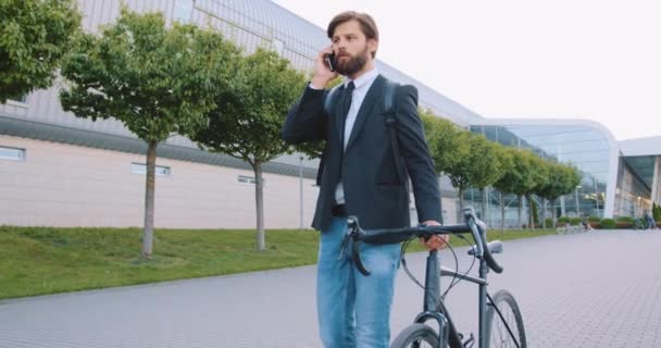 Retrato de bom temperamento confiante sério homem barbudo moderno em roupas elegantes que descendo com sua bicicleta na rua de paralelepípedos e falando no smartphone — Vídeo de Stock