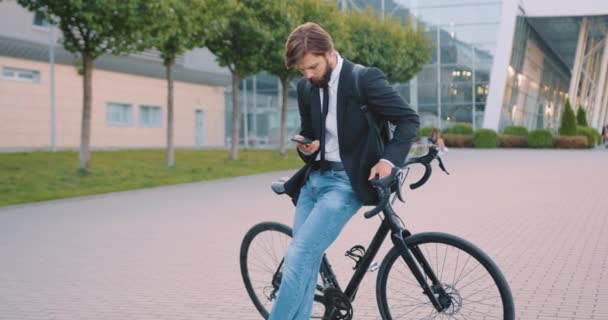 Schöne selbstbewusste positive junge bärtige Kerl, der neben seinem Fahrrad mit dem Handy auf dem Flughafen der Stadt Hintergrund steht — Stockvideo