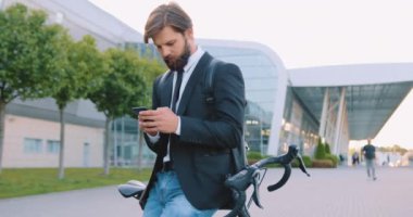 Modern havaalanı arka planında bisikletinin yanında duran yakışıklı, kendine güvenen, stil sahibi, genç sakallı adamın ön görüntüsü.