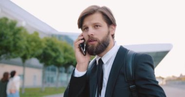 Güzel görünümlü, kendine güvenen, 30 yaşında, sakallı bir ofis yöneticisi. Şehir çağının modern binasının arka planında duruyor ve olumlu bir telefon konuşması yapıyor.