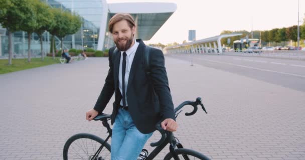 Hezký úsměv moderní mladý vousatý muž ve stylovém oblečení pózuje na kole uprostřed pěší stezky na impozantní krásné město letiště pozadí nebo jiné budovy — Stock video