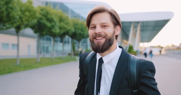 Sympatický veselý sebevědomý mladý muž s dobře udržovaným vousem ve stylovém oblečení s batohem pózující na kameru s upřímným úsměvem na městském dlážděné ulici pozadí — Stock video