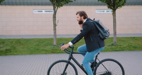 Tampilan samping dari manajer kantor muda berjanggut tampan dengan hati-hati dengan pakaian bergaya yang naik sepeda secara simultan berbicara di telepon — Stok Video