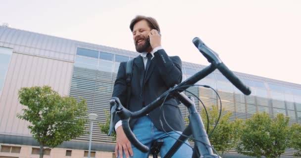 매력적 이고 명랑 한 젊은 남성 사무실 관리인 수염을 하고 현대의 큰 건물 근처에 있는 자신의 자전거에 앉아 긍정적 인 전화 대화를 즐기는 모습 — 비디오