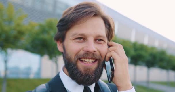 Retrato de atractivo joven feliz satisfecho con barba bien arreglada que disfruta de su conversación telefónica positiva en el fondo del edificio de la gran ciudad — Vídeo de stock