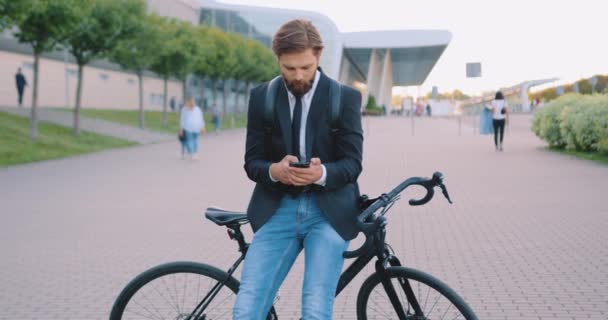 Вид спереду задоволений успішним молодим бородатим офісним працівником, який сидить на своєму велосипеді і друкує повідомлення по телефону на вражаючому міському будівельному тлі — стокове відео
