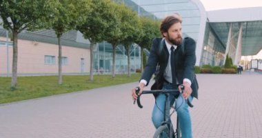 Sırt çantalı, resmi giysileri olan, hoş, genç, sakallı bir iş adamı büyük panoramik binanın yakınındaki kaldırım kenarındaki ofisine bisikletle gidiyor.