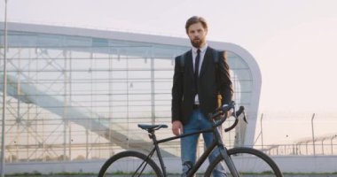 Yakışıklı, ciddi, 30 yaşında, sakallı bir ofis çalışanı. Gündüz vakti, büyük cam inşaat arka planında, bisikletinin yanında poz veriyor.