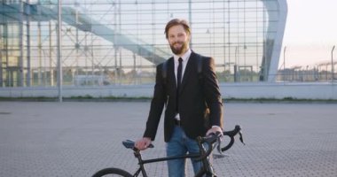 Yakışıklı, güler yüzlü, iş elbiseli, sakallı, modern cam bina arka planında bisikletiyle kameraya poz veren genç bir adam.