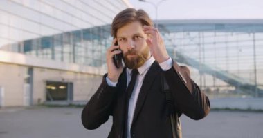 Şehrin modern cam binasının yakınında telefonla konuşan yakışıklı, mutlu, kendine güvenen, sakallı bir ofis çalışanının ön görüntüsü.