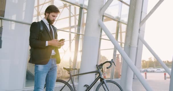 Красивий впевнений цілеспрямований молодий бородатий чоловік стоїть біля скляної стіни зі своїм велосипедом і використовує телефон для запису повідомлення, вид спереду — стокове відео