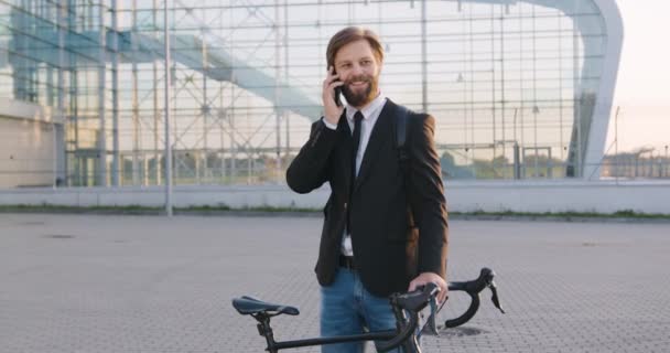 Gros plan de bonne humeur heureux satisfait barbu gars en vêtements formels qui debout près de son vélo et profiter de la conversation téléphonique sur fond de grand bâtiment en verre — Video