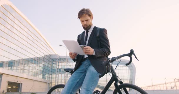 Вид спереду красивий концентрований зайнятий бородатий офісний менеджер, який стоїть біля свого мотоцикла біля великої міської будівлі та аналізує бізнес-документи — стокове відео