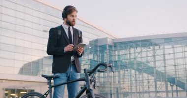 Modern şehir binasının arka planında motorunun yanında duran ve tablet bilgisayar kullanan kulaklıklı yakışıklı, konsantre genç iş adamının portresi.