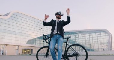 Sanal gerçeklik kulaklığıyla bisikletinin yanında duran çekici, kendine güvenen, genç, sakallı iş adamı ve kentsel cam bina arka planı üzerine hayali bir ekran üzerinde çalışıyor.