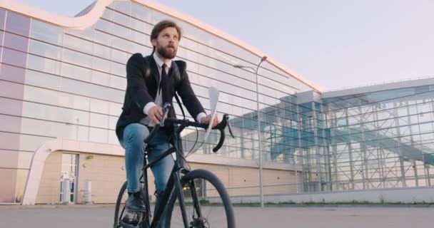 在现代城市玻璃建筑附近骑自行车，并将商业票据扔在头上的年轻、快乐、满脸胡须的生意人的眼前 — 图库视频影像