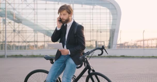 화려 한 턱수염을 한 30 세의 유능하고 자신감 있는 사무원이 대도시 유리 건물 주변에 서서 자전거 옆에 서 있는 동안 전화로 사업 문서에 대해 이야기 한다. — 비디오